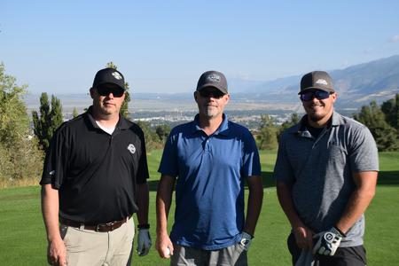 Three men standing on a golf course with mountains in the background.